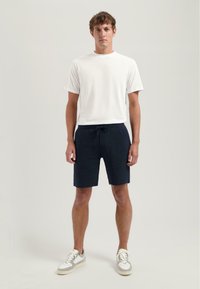 Tricou alb din bumbac, asortat cu pantaloni scurți texturat de culoare bleumarin, cu un brâu elastic și buzunare laterale, purtat cu adidași gri și albi.