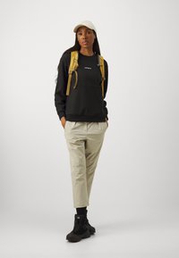 Black sweatshirt with ribbed cuffs, beige baggy trousers, black hiking boots, and a light-colored cap. Features a yellow backpack with straps.