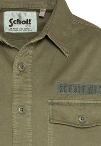 Olive green cotton shirt with a pointed collar, five-button placket, chest pocket with button, and embroidered logo. Soft texture, durable fabric.