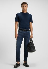 Marinblå kortärmad skjorta, mörkblå jeans, svart läderbälte, svarta loafers och en svart läderväska. Klassisk, figurnära siluett.