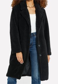 Manteau noir à texture polaire avec col à revers, fermetures à boutons et poches latérales. Porté sur un haut blanc et un jean bleu.