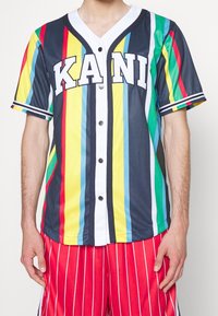 Meerlagig gestreept honkbalshirt met blauw, geel, rood en groen, met witte accenten en "KANI" in grote letters aan de voorkant.