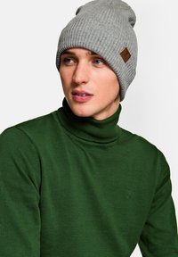 Szara, prążkowana czapka beanie z podwiniętym mankietem i małą brązową naszywką z logo, noszona z ciemnozielonym swetrem z golfem na jasnym tle.