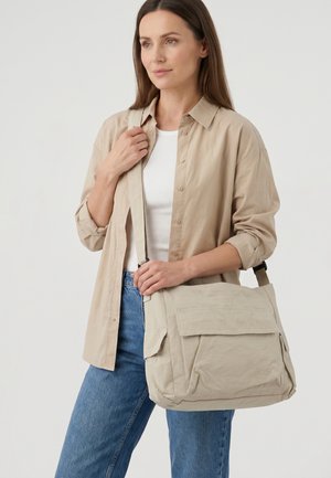 Woman in beige shirt and blue jeans holding a large beige shoulder bag with multiple pockets against a plain background.
