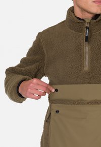 Pullover in pile verde oliva con colletto alto, chiusura frontale con zip e tasca inferiore con bottoni a pressione. Texture morbida e design caldo.