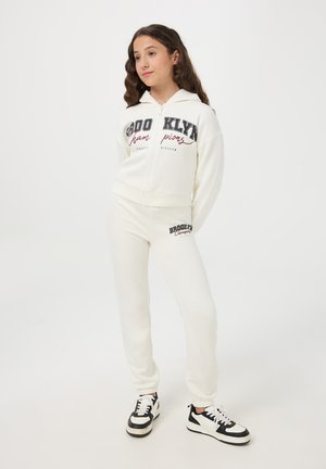 Felpa bianca con zip e pantaloni abbinati, con la scritta "BROOKLYN" in nero e maroon. Tessuto morbido e vestibilità rilassata, abbinati a sneakers in bianco e nero.