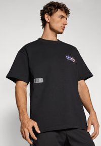 Svart oversized T-skjorte laget av bomull, med grafikk på forsiden i lilla og en transparent etikettakcent på nedre venstre hjørne.