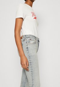Camiseta blanca con texto gráfico rojo, combinada con unos jeans de mezclilla de lavado claro. Los jeans tienen un ligero desvanecimiento y un diseño clásico de cinco bolsillos.