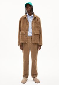 Bruine corduroy jas met bijpassende broek, blauwe shirt eronder en groene pet. Lichtbruine instap schoenen. De textuur is geribbeld.