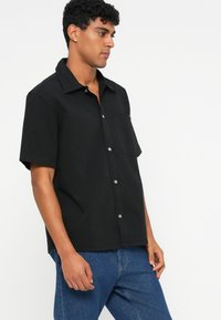 Črna kratkotrična srajca iz bombaža z gumbom spredaj, enim žepom na prsih in subtilnim sijajem srebrnih gumbov.