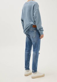 Persona che indossa un ampio felpa azzurro chiaro, jeans blu strappati, calze bianche e sneakers bianche su uno sfondo semplice.