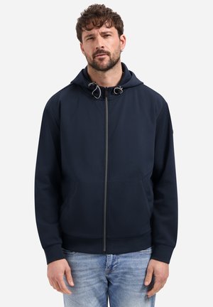 Mann mit kurzem lockigem Haar, der einen marineblauen Kapuzenpullover mit Reißverschluss und hellblaue Jeans trägt, steht vor einem einfachen weißen Hintergrund.