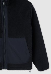 Giacca in pile nera, con zip frontale, tessuto strutturato e pannelli lisci a contrasto. Include due grandi tasche frontali.