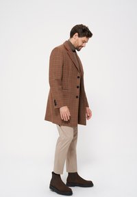 Brązowy płaszcz w kratę z czarnymi guzikami, z klapami i bocznymi kieszeniami, w połączeniu z beżowymi spodniami i brązowymi butami typu chelsea.