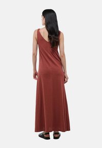 Robe maxi de couleur rouille, avec un dos nu, sans manches, fabriquée en tissu doux avec une silhouette fluide, associée à des sandales noires.