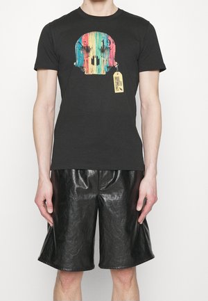 Sort bomuld t-shirt med et farverigt kraniedesign på forsiden og en gul mærkat. Kombineret med skinnende sorte lædershorts.