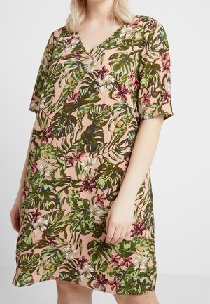 Kobieta ubrana w sukienkę z krótkim rękawem i dekoltem w kształcie litery V, na różowym tle z zielonym, białym i czerwonym tropikalnym wzorem liści i kwiatów.