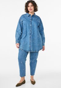Denim set featuring a long shirt with button closure and a curved hem, paired with matching tapered pants, both adorned with floral embroidery.