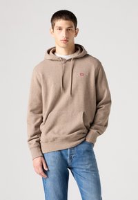 Felpa beige con cappuccio e finitura testurizzata, tasca a marsupio frontale e logo rosso sul petto, abbinata a jeans azzurri chiari.
