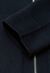 Dettaglio di giacca in lana nera con polsino spesso, texture liscia e cerniera argentata con dentini visibili lungo la cucitura frontale.