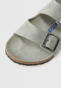 Γκρίζα σουέτ σανδάλι με δύο φαρδιές λωρίδες, μεταλλικό κούμπωμα και υφασμένο πάτο με καφέ φελλό και μαύρη εξωτερική σόλα από καουτσούκ.