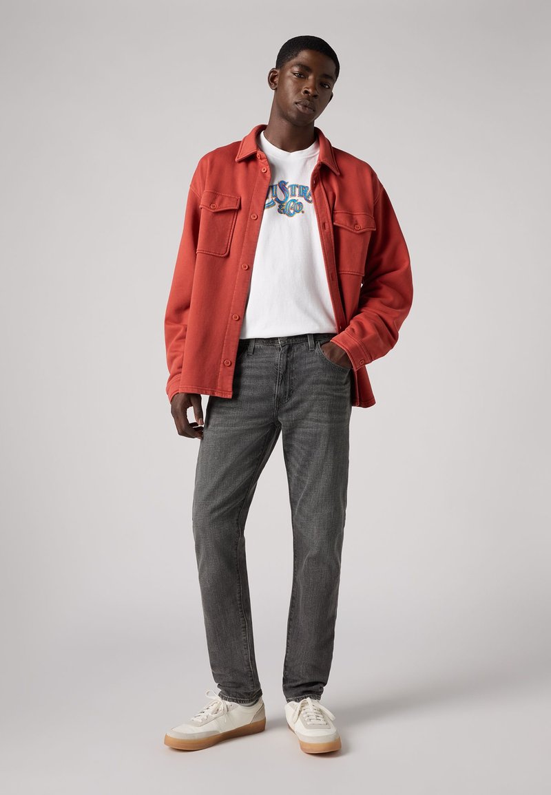Veste rouge à boutons, t-shirt graphique blanc, jeans gris et sneakers blanches avec semelle en caoutchouc. Le mannequin se tient avec les bras détendus le long du corps.