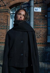 Cappotto di lana nera con un ampio colletto drappeggiato, due tasche frontali e due bottoni. Il tessuto ha una sottile trama, posizionato davanti a un muro di mattoni.