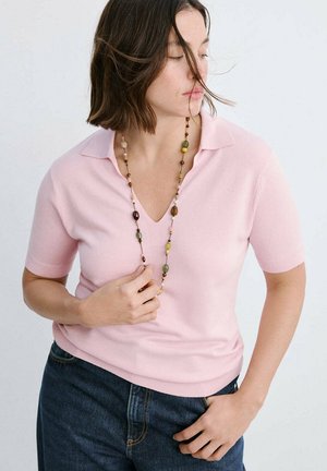 Femme aux cheveux bruns mi-longs porte un haut rose à manches courtes avec col, un jean bleu et un long collier de perles, regardant vers le bas à gauche.