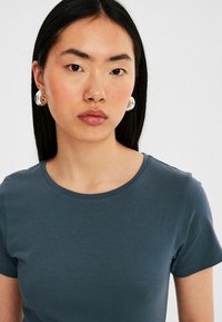Mörkblå t-shirt med rund halsringning, kort ärm, tillverkad av ett glatt tyg. Modellen bär stora runda klara örhängen. Neutral bakgrund.