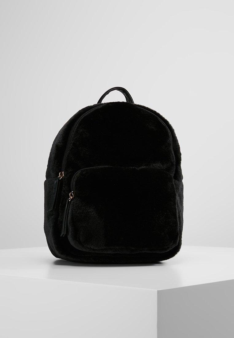 Sac à dos en tissu peluche noir avec poche zippée devant et poignée supérieure, présenté sur une surface blanche sur fond gris clair.