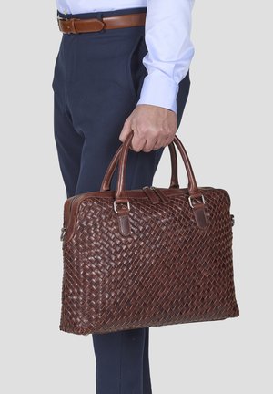 Borsa in pelle intrecciata marrone con doppio manico, chiusura con zip e hardware argentato, tenuta da una persona che indossa pantaloni blu navy e una camicia azzurra.