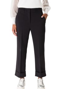 Femme portant un pantalon noir droit avec ourlets, un chemisier blanc à manches en dentelle et des baskets argentées, debout avec une main dans la poche.