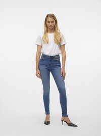 Ženska z dolgimi svetlimi lasmi, oblečena v belo majico, modre skinny jeans z visokim pasom in črne mule s koničastim prstom ter visokimi petami, stoji obrnjena naprej.