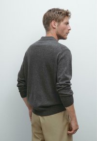 Dunkelgrauer, gestrickter Pullover mit rundem Halsausschnitt, Rippbündchen und Saum. Getragen mit hellbeigen Hosen, die eine figurbetonte Silhouette zeigen.