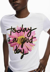 Witte katoenen t-shirt met een grote roze bloemenafbeelding en geborduurde tekst die zegt: "Vandaag is een goede dag" in het zwart. Korte mouwen.