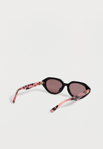Lunettes de soleil avec des montures angulaires noires et des branches roses à motif écaille reposant sur une surface blanche.