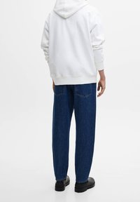 Sudadera blanca con capucha, puños y dobladillo acanalados, combinada con jeans de mezclilla oscuros de corte holgado con bolsillos traseros. Calzado negro visible.
