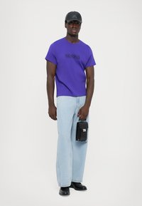 Μωβ κοντομάνικο μπλουζάκι με εκτύπωση «Ο ΛΑΟΣ», ανοιχτό μπλε ίσια παντελόνια τζην, μαύρη τσάντα χεριού και μαύρα παπούτσια. Το μοντέλο στέκεται μπροστά σε λευκό φόντο.