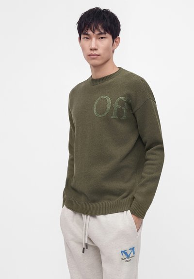 Jeune homme aux cheveux noirs portant un pull vert olive avec le texte « "Off" » et un pantalon de survêtement gris clair avec un logo flèche bleu, debout devant un fond blanc.