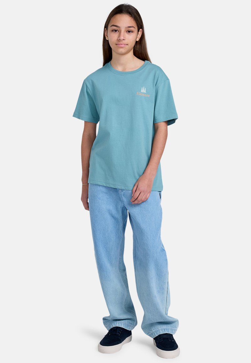Camiseta de algodón de color azul claro con mangas cortas y cuello redondo, que presenta un pequeño logo naranja. Se lleva con pantalones de mezclilla azul claro y zapatos oscuros.