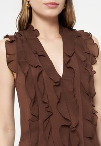 Femme portant un chemisier marron sans manches avec des détails de volants superposés le long du décolleté en V et des épaules sur un fond uni.