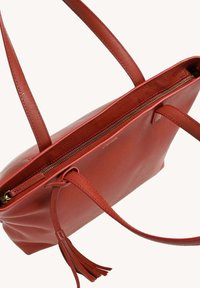 Sac fourre-tout en cuir rouge avec un dessus zippé, deux longues poignées et un détail de gland. Présente une texture lisse et une forme structurée.