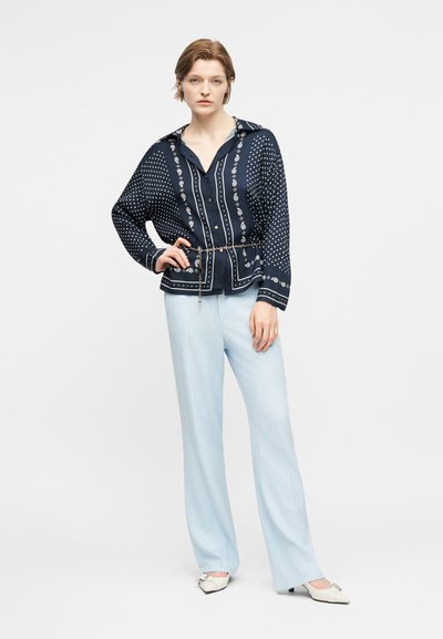 Γυναίκα που στέκεται με το χέρι στη μέση, φοράει μπλε πουκάμισο με μοτίβο και κουμπιά, ανοιχτόμπλε παντελόνι φαρδύ και λευκά παπούτσια με τακούνι.