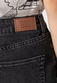 Černá denimová sukně s texturovaným povrchem. Na pásku je hnědá kožená nášivka s označením "BDG" a viditelné detaily šití.