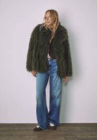 Zelena, puhasta prevelika jakna z teksturiranim zaključkom, nošena preko temnega topa in modrih širokih kavbojk. Postavljena na nevtralno ozadje.