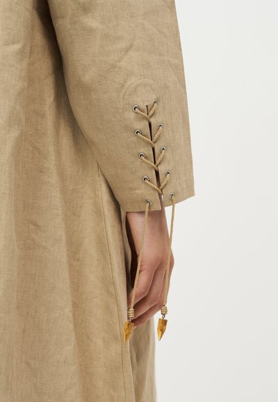 Gros plan d'une manche en lin beige avec un laçage décoratif en cordelette croisée et des extrémités de cordelette de couleur ambre suspendues à côté d'une main.
