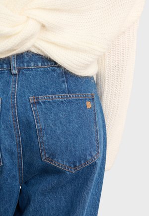 High-waisted blue jeans with two back pockets, orange stitching, and a subtle logo patch on the right pocket. Soft white knitted sweater visible above.