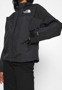 Svart vattentät jacka med dragkedja fram, huva, justerbara ärmslut och The North Face-logotyp på bröstet. Slät textur och figursydd design.