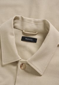 Smėlio spalvos marškiniai su apykakle, vienu smėlio spalvos mygtuku ir vidinėje iškirptės pusėje prisiūta juoda audinio etiketė su užrašu „Matinique“.