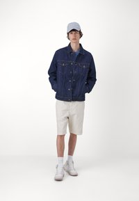 Denim jakna u tamnoplavoj boji s dva džepa na grudima, zatvaranje na dugmad i opušteni kroj. Uparena s bež šortsevim i bijelim tenisicama.